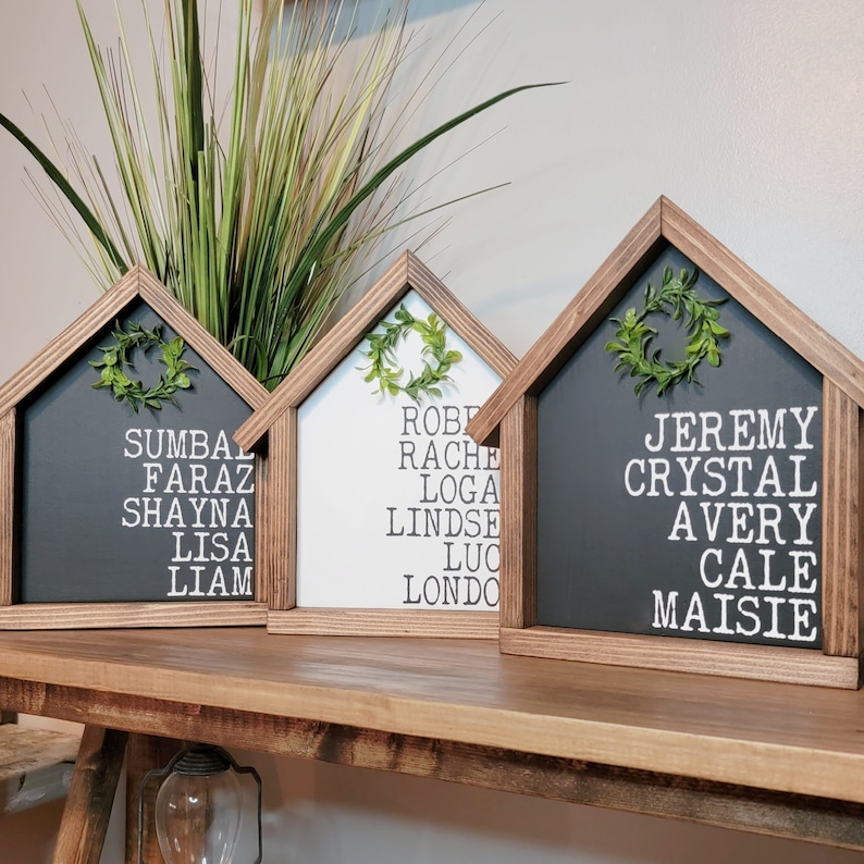 May include: Three wooden house-shaped signs with black, white, and grey backgrounds. Each sign has a wreath and a list of names in white lettering. The names on the signs are: Sumbaz, Faraz, Shayna, Lisa, Liam, Robe, Rache, Loga, Lindse, Luc, Londo, Jeremy, Crystal, Avery, Cale, Maisie.
