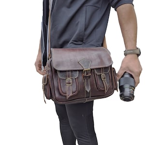 Peut inclure: Un sac à bandoulière en cuir marron avec plusieurs poches et une fermeture à boucle. Le sac est porté sur l'épaule d'une personne vêtue d'un pantalon noir et d'une veste gris foncé.