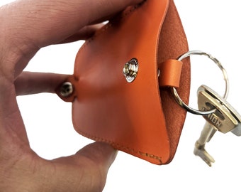 May include: Orange leather key holder with a metal snap closure and a silver key ring. The key holder is open and a silver key is attached to the ring.