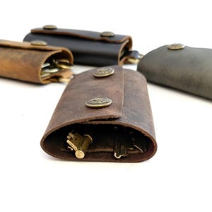 May include: Four leather key holders in different colours, brown, dark brown, grey, and black. Each holder has a flap with a brass button closure. The holders are filled with keys.