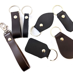 May include: Six different leather keychains with metal rings. The keychains are in various shapes and sizes, including a rectangle, a leaf, and a teardrop. Some of the keychains have a metal clasp.