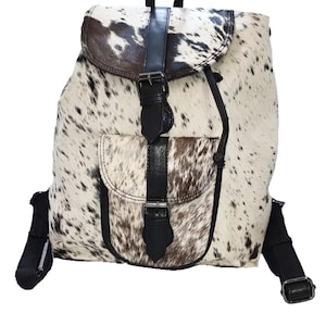 May include: A cowhide backpack with a spotted pattern in shades of brown and white. The bag features black leather accents, including straps, a flap closure, and a front pocket. Adjustable shoulder straps and a top handle are also visible.