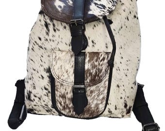 May include: A cowhide backpack with a spotted pattern in shades of brown and white. The bag features black leather accents, including straps, a flap closure, and a front pocket. Adjustable shoulder straps and a top handle are also visible.