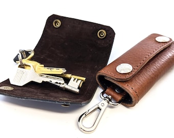 May include: A brown leather key holder with a silver snap closure and a silver hook. The key holder is open and has a set of keys inside.