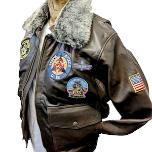 Op de afbeelding: Een bruine leren bomberjack met een nep bontkraag en meerdere patches, waaronder "Strike Attack", "Top Gun" en "Tomcat".