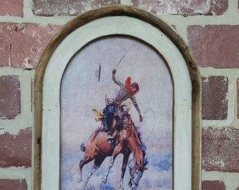 May include: A framed print of a cowboy riding a bucking horse. The cowboy is wearing a red bandana and a hat. The horse is brown with white markings. The print is set against a light blue background.