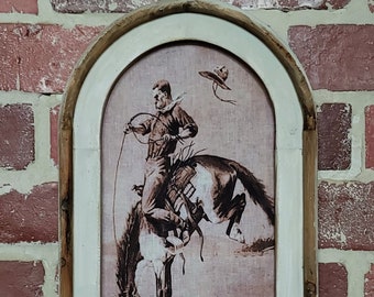 May include: A framed print of a cowboy riding a bucking horse. The print is in shades of brown and white and has a textured background. The frame is a rustic wood with a white painted finish.
