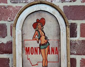 May include: A framed print of a woman in a red cowboy hat and a turquoise outfit standing in front of the state of Montana. The print is on a canvas background with a white frame.