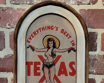 May include: A framed vintage-style sign with a woman in a cowboy hat and boots. The sign says "Everything's Best in Texas" in red letters.