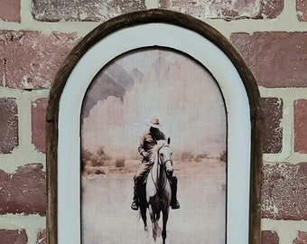 May include: Arched wall art featuring a cowboy on horseback in a serene landscape. The artwork is framed in a rustic wooden frame with a white inner border. The cowboy is wearing a hat and riding a white horse through water, with a reflection below.