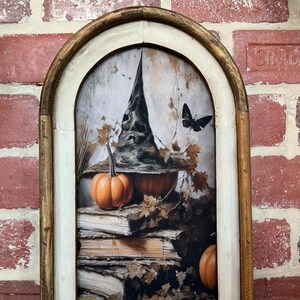 May include: A framed print of a witch's hat with pumpkins and books. The hat is black with a pointed top and a wide brim. The pumpkins are orange and the books are stacked on top of each other. The background is a light brown color.