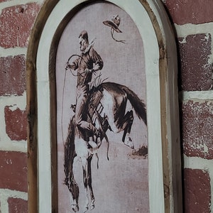 May include: A framed sepia-toned print of a cowboy riding a bucking horse. The frame is made of wood with a whitewashed finish and has an arched top.