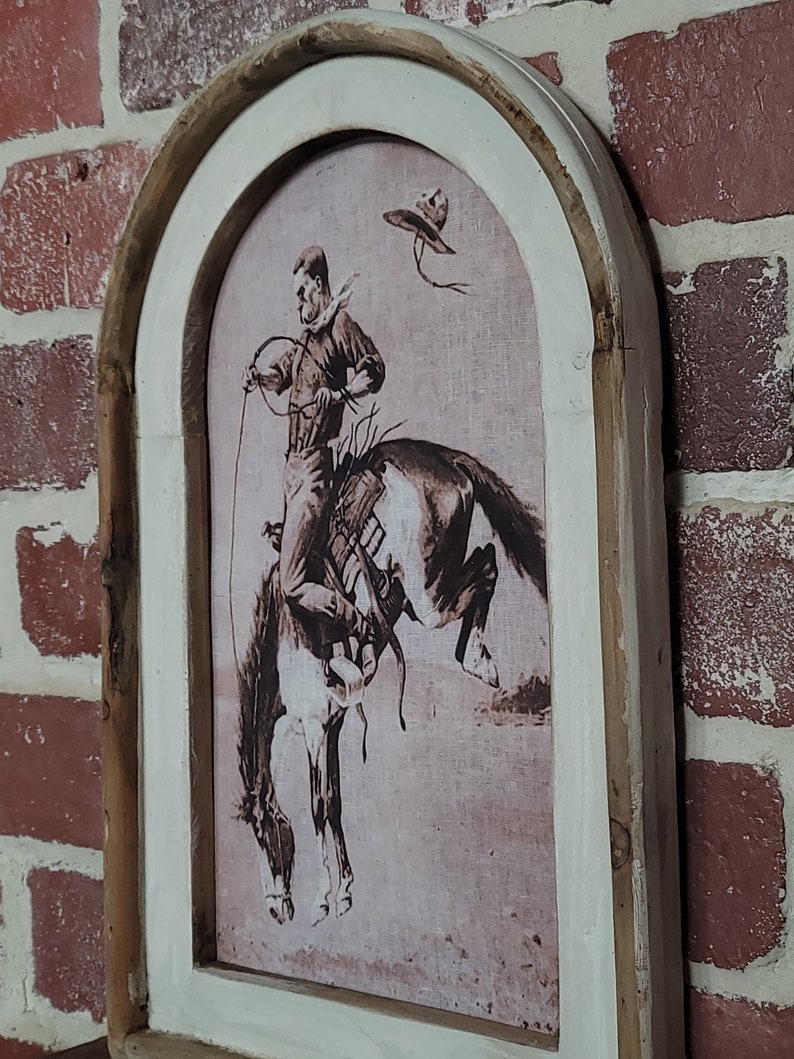 May include: A framed sepia-toned print of a cowboy riding a bucking horse. The frame is made of wood with a whitewashed finish and has an arched top.