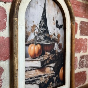 May include: A framed print of a witch's hat with a pumpkin and books. The hat is black with a pointed top and a wide brim. The pumpkin is orange and sits on top of a stack of old books. The background is a faded, vintage-style image of leaves and branches.