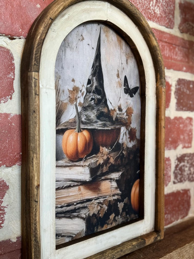 May include: A framed print of a witch's hat with a pumpkin and books. The hat is black with a pointed top and a wide brim. The pumpkin is orange and sits on top of a stack of old books. The background is a faded, vintage-style image of leaves and branches.