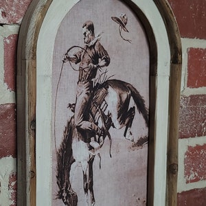 May include: A framed print of a cowboy riding a horse. The print is in black and white and has a vintage feel. The frame is made of wood and has a distressed white finish.