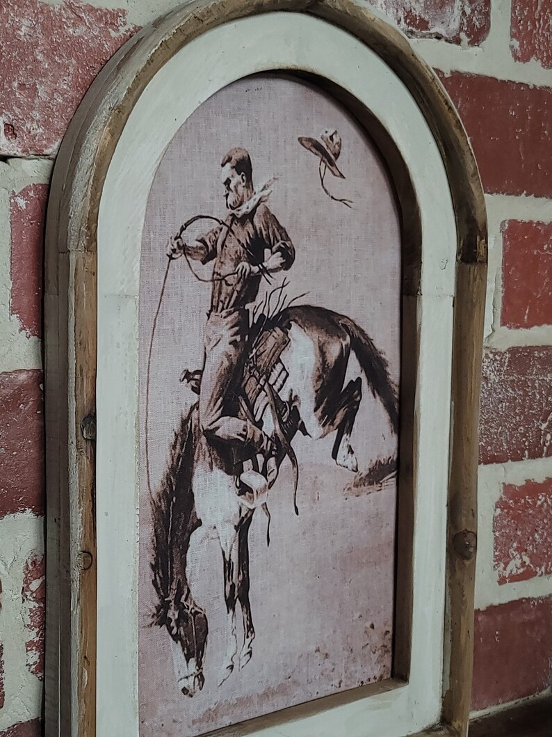 May include: A framed print of a cowboy riding a horse. The print is in black and white and has a vintage feel. The frame is made of wood and has a distressed white finish.