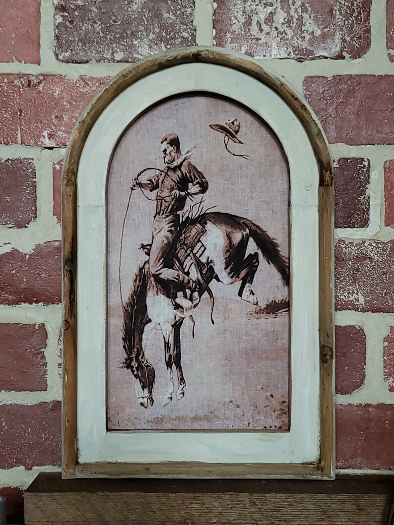 May include: A framed print of a cowboy riding a bucking horse. The print is in shades of brown and white and has a textured background. The frame is a rustic wood with a white painted finish.