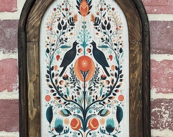 May include: A framed artwork featuring a symmetrical design with two black birds facing each other. The artwork includes orange, teal, and white floral elements. The frame is made of dark brown wood and has an arched top.