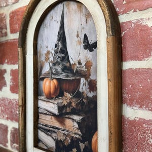 May include: A framed print with a witch's hat, pumpkins, and a butterfly. The print is in a rustic wooden frame with a white inner border. The background is a dark, textured surface.