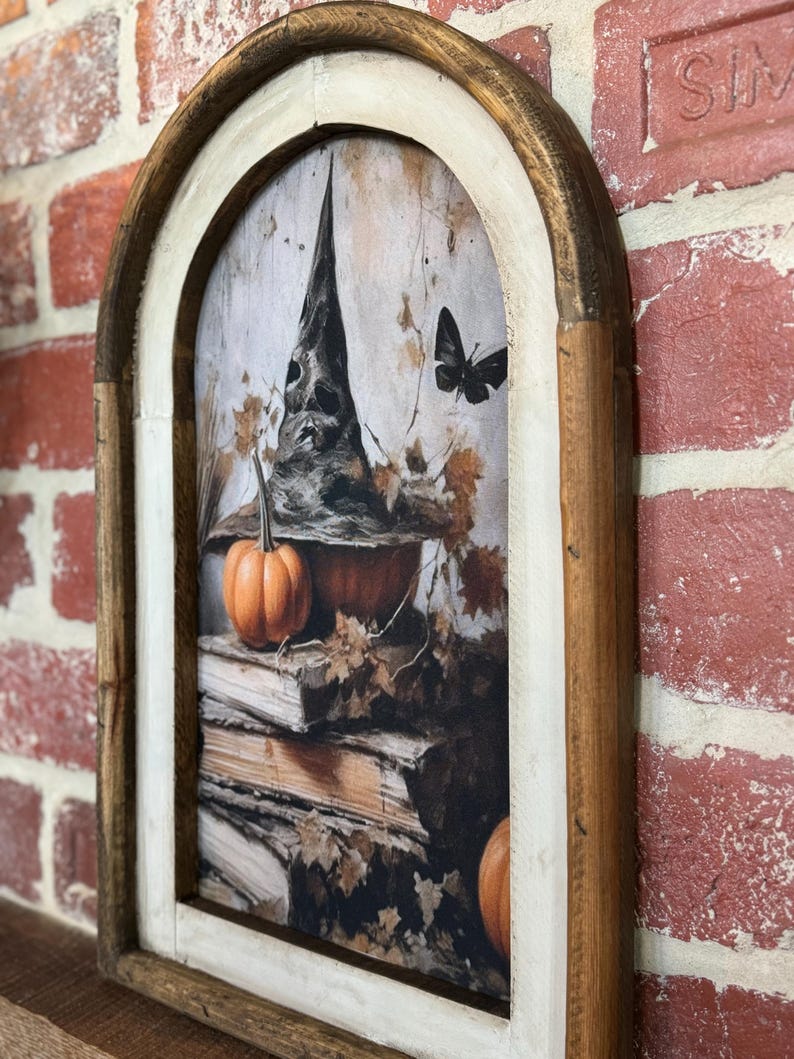 May include: A framed print with a witch's hat, pumpkins, and a butterfly. The print is in a rustic wooden frame with a white inner border. The background is a dark, textured surface.