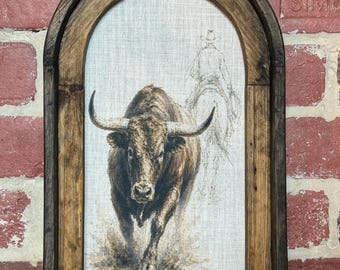 May include: An arched wall art piece featuring a dark brown wooden frame surrounding a canvas print. The print depicts a brown bull in the foreground and a faint, sketched outline of a cowboy on horseback in the background.