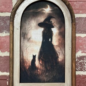 May include: Arched framed canvas art featuring a witch silhouette in a black dress and hat, standing in a field with a black cat. The background shows a moonlit sky. The frame is made of wood with a distressed finish.