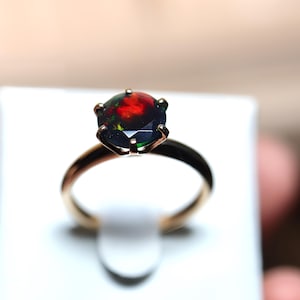 Puede incluir: Un anillo de oro con una gran piedra preciosa de ópalo negro redondo con destellos rojos, verdes y amarillos vibrantes. El ópalo está engastado en un engaste de seis puntas, y la banda es de color oro pulido. El anillo se muestra sobre una superficie blanca.