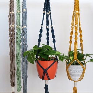 Puede incluir: Cuatro macramés para colgar plantas con cuentas de madera, cada uno en un color diferente: marrón, verde, azul y amarillo. Los macramés están colgados de una barra verde. Dos de los macramés tienen plantas en macetas.