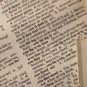 May include: A close-up shot of old dictionary pages, featuring definitions for words such as "confidence" and "confide." The paper has a warm, aged appearance, and the text is printed in a traditional serif typeface.