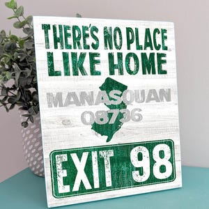May include: A rectangular sign with a white wood grain background featuring the green text THERE'S NO PLACE LIKE HOME, a green silhouette of New Jersey, the grey text MANASQUAN 08736, and a green highway sign graphic reading EXIT 98.