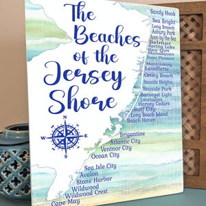 May include: A framed print featuring a map of the Jersey Shore with a watercolor effect. The title "The Beaches of the Jersey Shore" is in elegant script. A compass rose is at the bottom left, and beach names are listed along the coast.