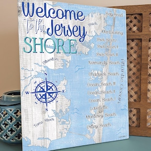Puede incluir: Un letrero de madera azul y blanco con un mapa de la costa de Jersey. El letrero dice "Bienvenido a la costa de Jersey" e incluye los nombres de varias ciudades y playas a lo largo de la costa. Una br&uacute;jula est&aacute; en el centro del mapa.