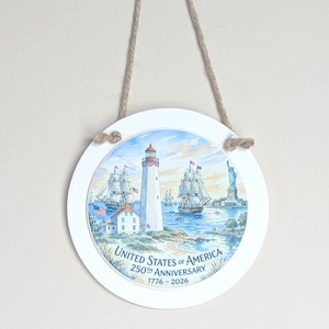 May include: A round, white hanging sign with a nautical scene featuring a lighthouse, sailing ships, and the Statue of Liberty. The sign commemorates the 250th anniversary of the United States of America, with text reading "United States of America 250th Anniversary 1776-2026".