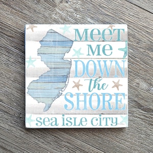 Puede incluir: Letrero cuadrado de madera con un dise&ntilde;o envejecido. El letrero presenta el contorno de Nueva Jersey en azul y blanco, con las palabras "Meet Me Down the Shore" y "Sea Isle City" en texto azul. Estrellas de mar decorativas dispersas.