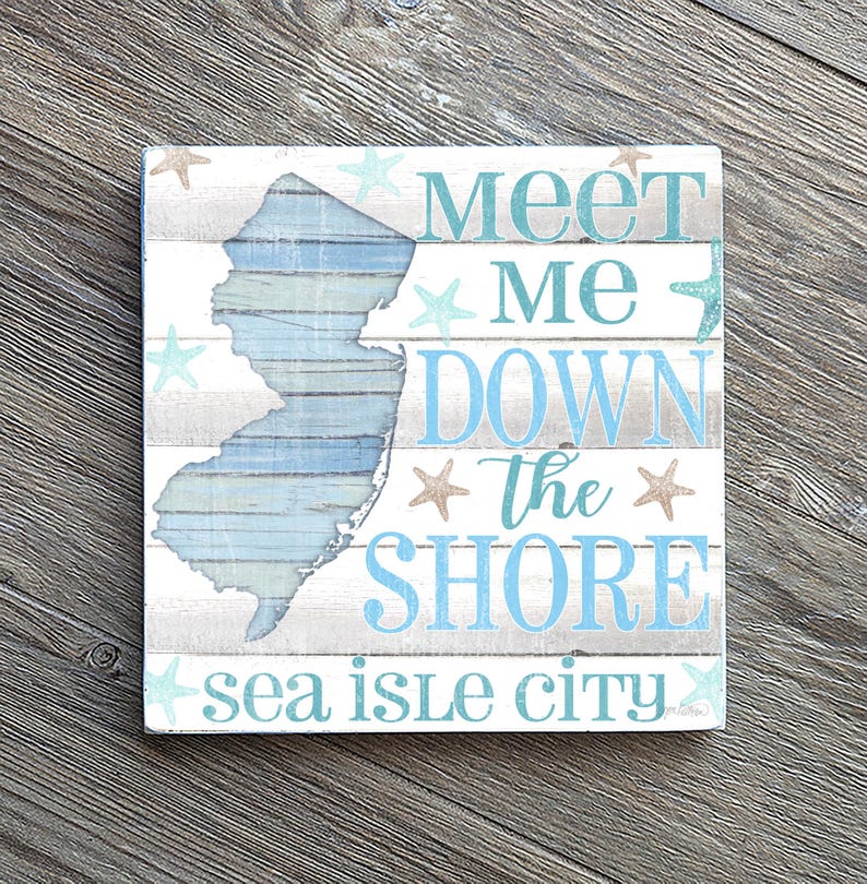 Puede incluir: Letrero cuadrado de madera con un dise&ntilde;o envejecido. El letrero presenta el contorno de Nueva Jersey en azul y blanco, con las palabras "Meet Me Down the Shore" y "Sea Isle City" en texto azul. Estrellas de mar decorativas dispersas.