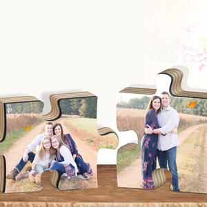 May include: Two wooden jigsaw pieces with photos of families. The left piece shows a family of four sitting on a path in a field. The right piece shows a couple standing on a path in a field.