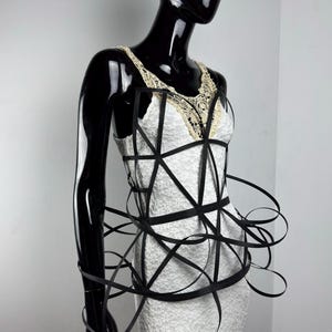 May include: A white lace dress with a black geometric design overlay. The dress features a cream-colored lace neckline and is displayed on a black mannequin. The black design wraps around the bodice and waist.