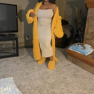 May include: A woman wearing a long, mustard yellow knitted cardigan with a matching pair of knitted slippers. The cardigan has a loose, oversized fit and the slippers are fluffy and comfortable.