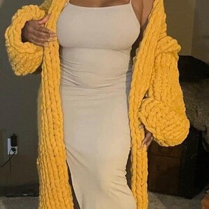 Pode incluir: Uma mulher vestindo um cardigã de malha grossa amarelo mostarda, chinelos combinando e um vestido bege. O cardigã tem um design longo e aberto com mangas grandes. O vestido é um estilo simples e justo.