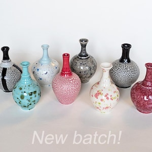 May include: A collection of eight small ceramic vases with narrow necks and bulbous bodies, featuring various glazes including crackle, speckled, and solid finishes in colours like black, white, pink, blue, and red, with the text New batch! below.
