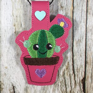 May include: A pink cactus-shaped keychain with a metal ring. The keychain features a green cactus in a brown pot, a light blue heart, and a purple flower. The cactus has a smiling face and is made of embroidered fabric.