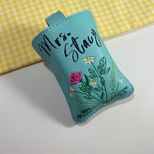 May include: A light blue key fob with the embroidered name "Mrs. Stacy" in black script. The fob features a floral design with pink roses, white daisies, and green leaves. A small loop is at the top.