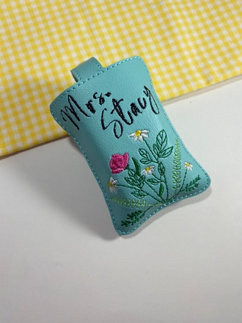 May include: A light blue key fob with the embroidered name "Mrs. Stacy" in black script. The fob features a floral design with pink roses, white daisies, and green leaves. A small loop is at the top.