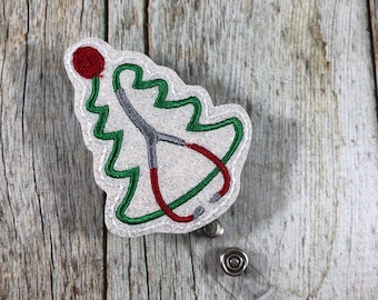 May include: A badge reel featuring a Christmas tree design with a stethoscope. The tree is outlined in green with a red ornament at the top. The stethoscope is red and gray. The badge reel has a clear plastic clip and a metal button.
