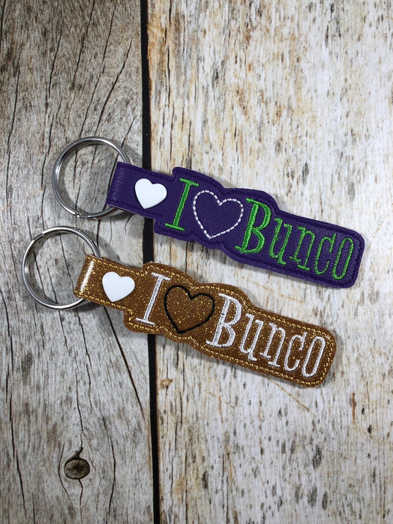 Może przedstawiać: Dwa breloczki do kluczy z napisem "I ❤️ Bunco". Jeden breloczek jest fioletowy z zielonymi i białymi akcentami, drugi jest złoty z brązowymi i białymi akcentami. Oba mają srebrny k&oacute;łko na klucze.