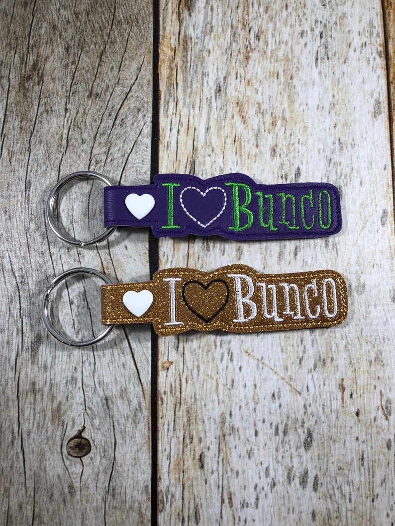 Może przedstawiać: Dwa breloki do kluczy z napisem "I Heart Bunco". Jeden jest fioletowy z zielonym tekstem i białym sercem, drugi jest złoty z białym tekstem i czarnym sercem. Oba mają srebrne k&oacute;łka na klucze.