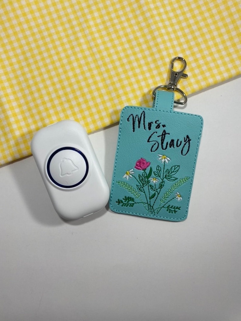 May include: A white doorbell with a blue bell icon next to a turquoise luggage tag. The tag is embroidered with a floral design and the name "Mrs. Stacy" in black script. A silver clasp is attached to the tag.