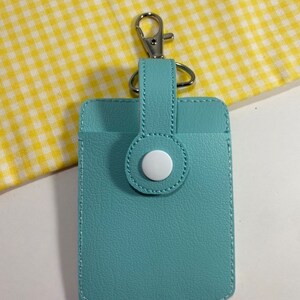 May include: A light blue cardholder with a silver-colored clasp and a white button closure. The cardholder is rectangular with a stitched border and a loop for the clasp.