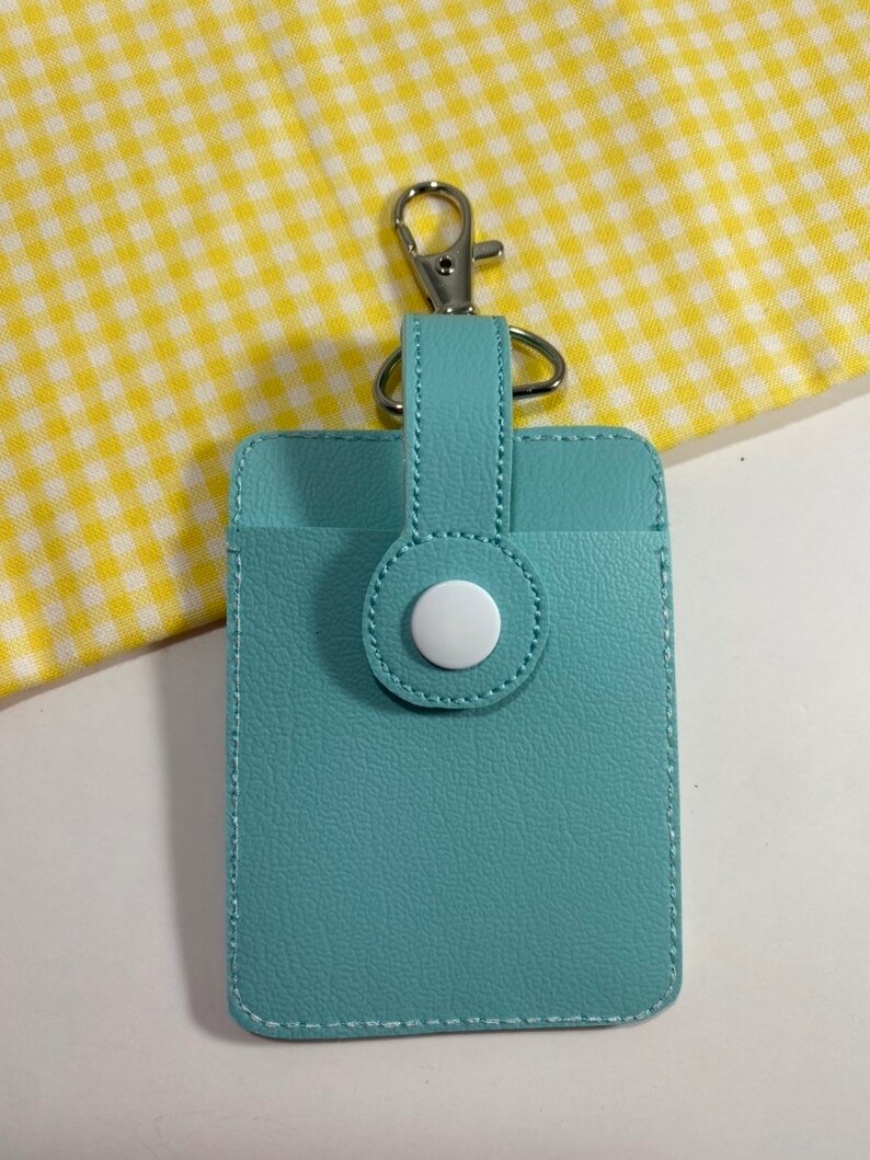 May include: A light blue cardholder with a silver-colored clasp and a white button closure. The cardholder is rectangular with a stitched border and a loop for the clasp.